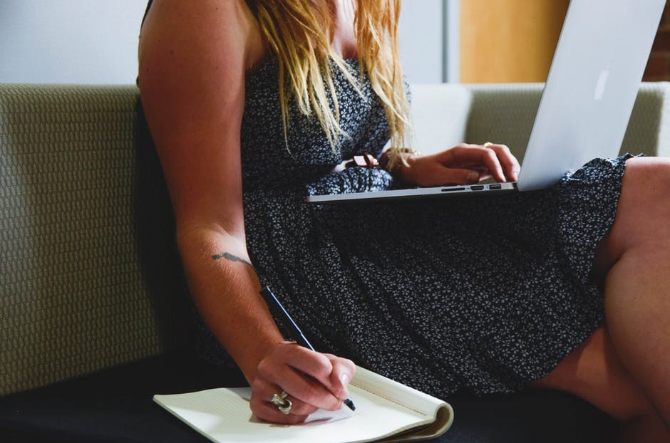 woman in dress typing at computer and writing in notebook